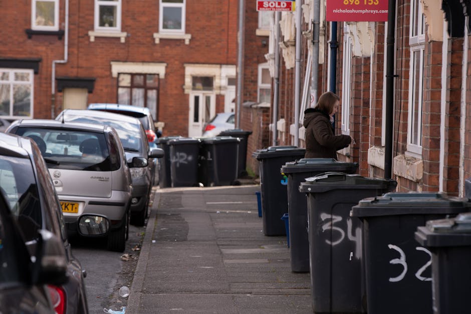 A row of large black wheelie bins with closed lids lined up along a paved residential sidewalk beside a brick building, which features multiple white-framed windows and a red sign indicating propertie